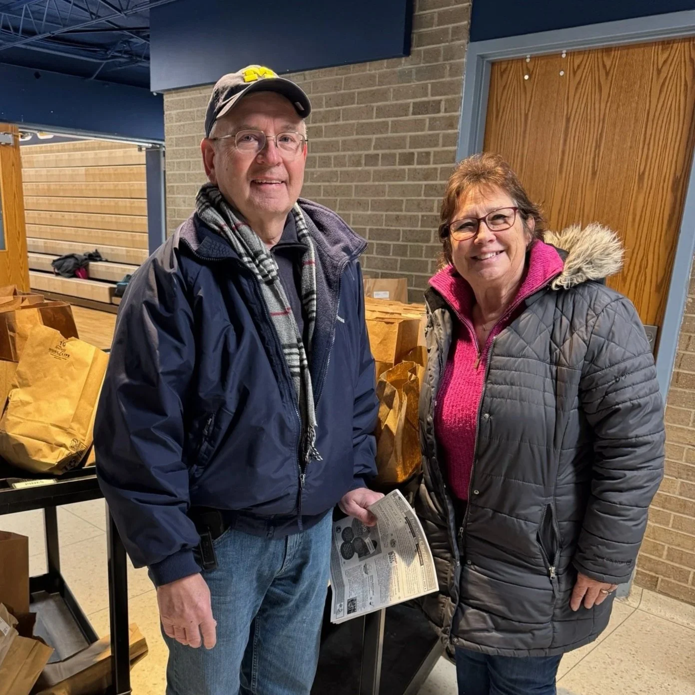 A smiling older man and woman stand indoors near a table with paper bags, both wearing jackets. The man is holding a flyer, and the woman wears glasses and a jacket with a fur-lined hood.