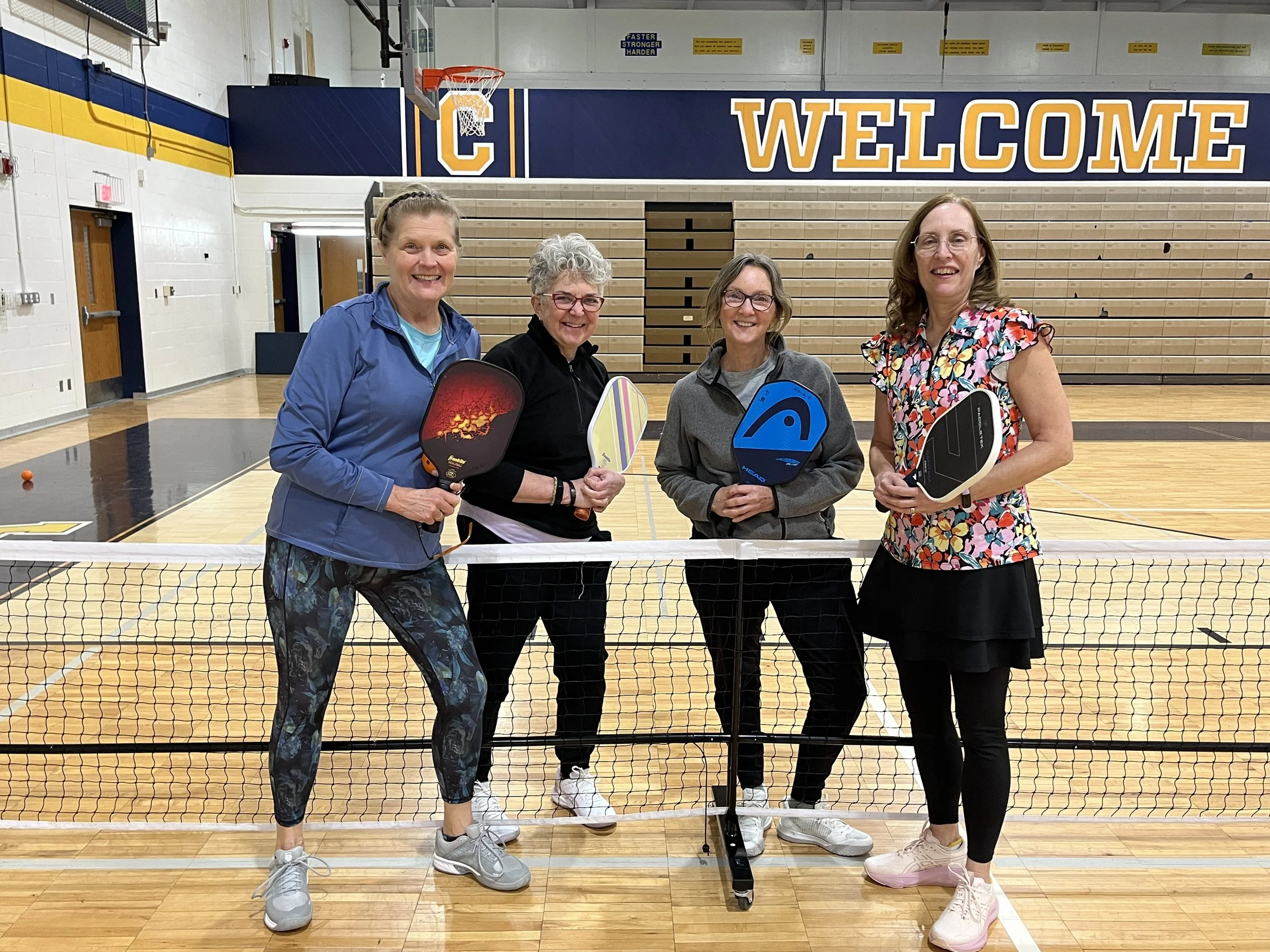 Four women standing on a gymnasium floor, holding pickleball paddles, behind a net, with a large yellow and blue 'WELCOME' sign on the wall in the background.