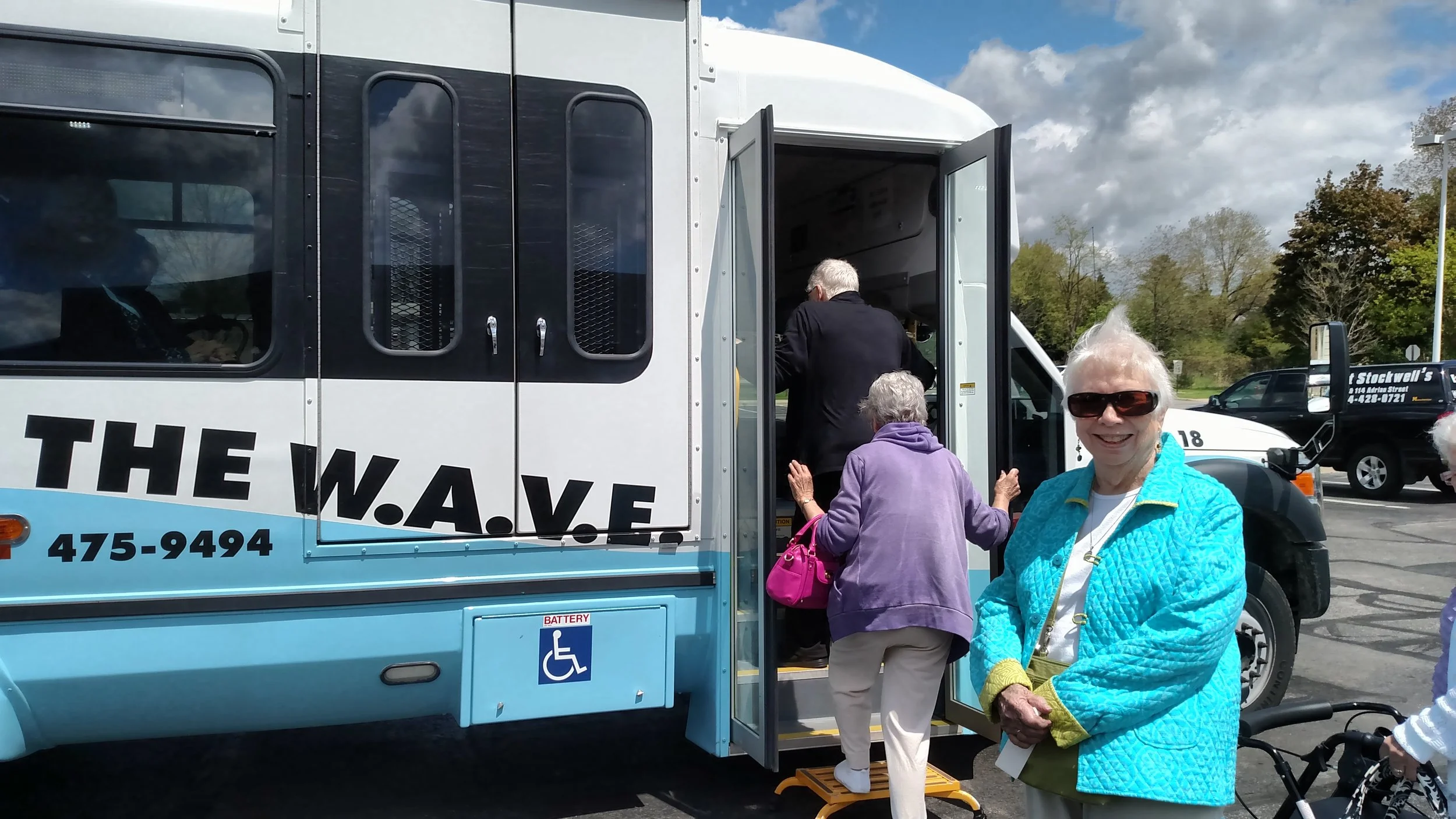 Older women boarding a bus with a woman smiling at the camera in the foreground, in a parking lot with trees and another vehicle in the background.
