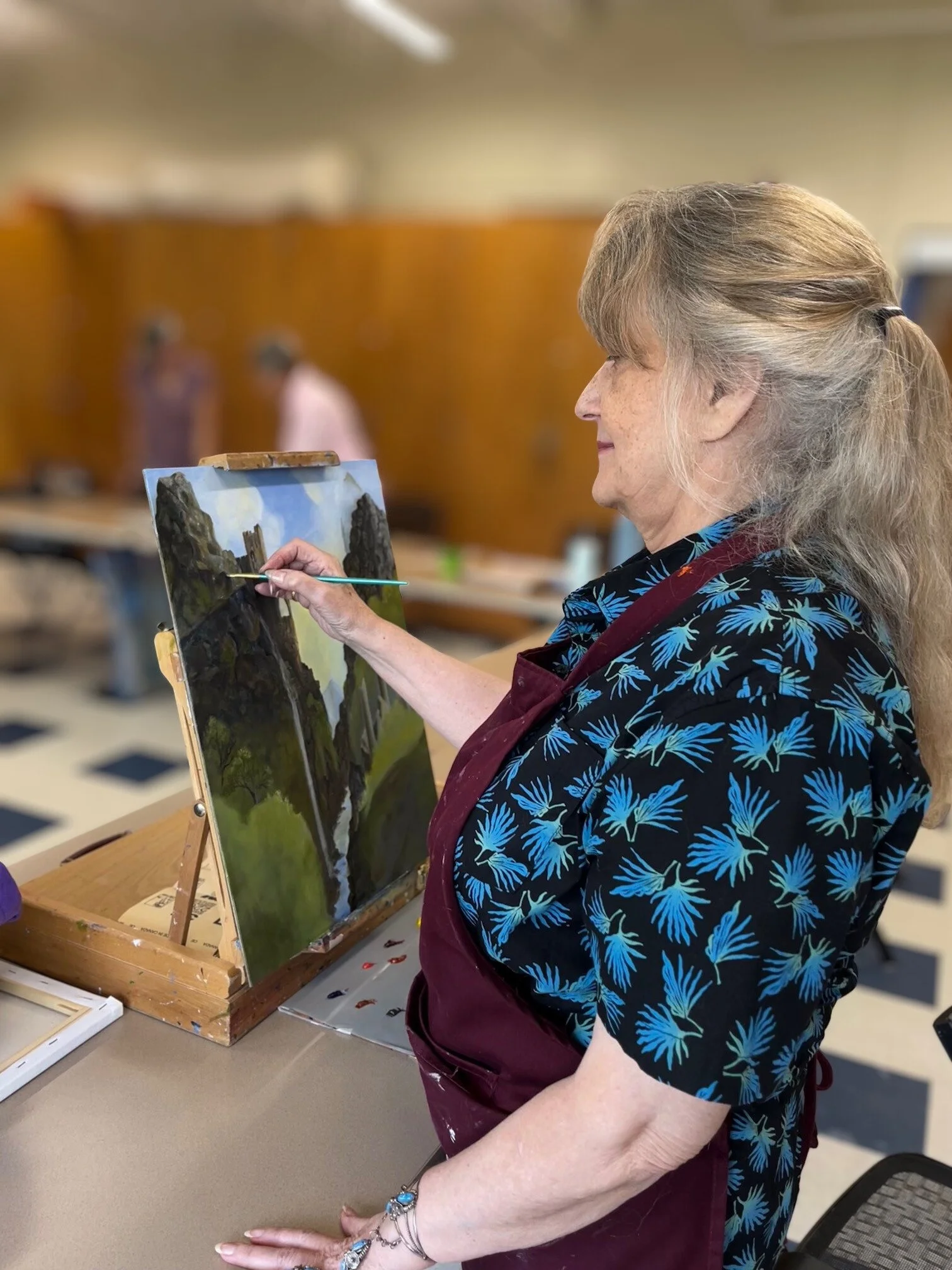 An elderly woman with gray hair painting a landscape on a small canvas at an art class. She is wearing a blue patterned shirt and a maroon apron. In the background, a blurred view of other people in the art studio.