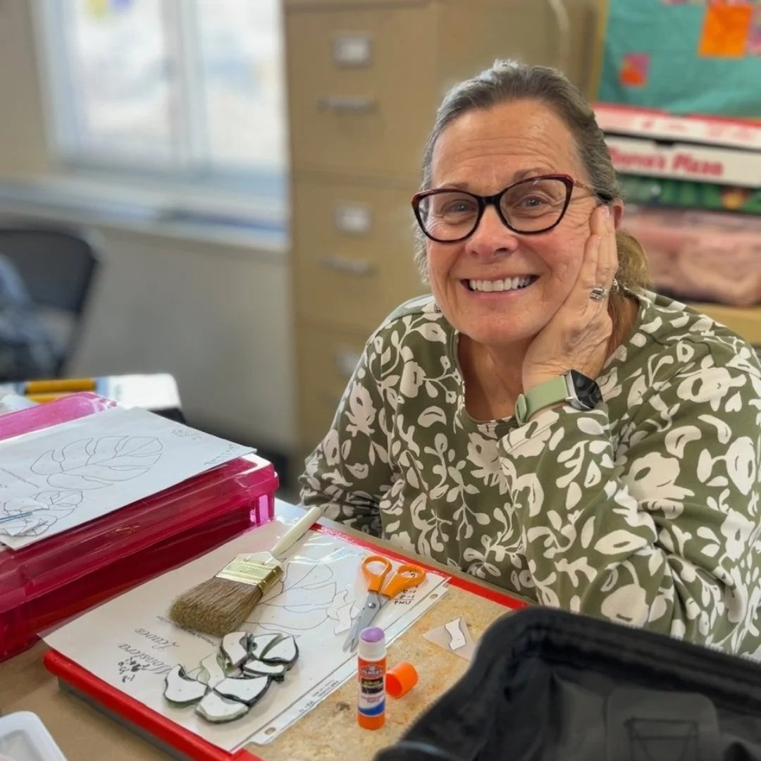 A woman wearing glasses and a patterned shirt, smiling at a table with arts and crafts supplies, including paper, scissors, glue, and a paintbrush. She is touching her face with her left hand and has a smartwatch on her wrist.