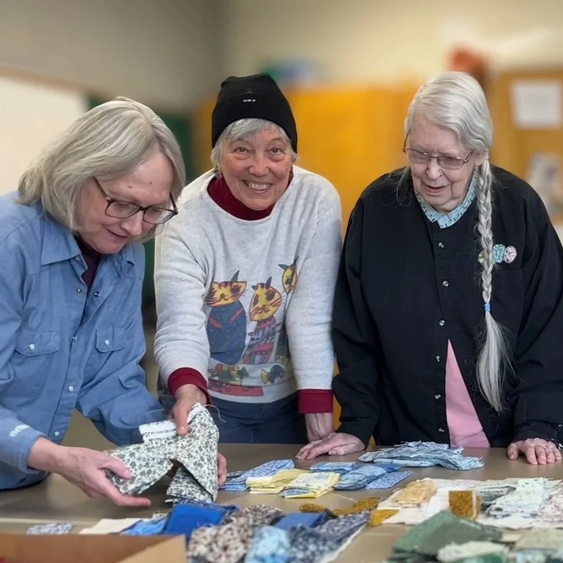 Three women examining fabric swatches on a table in a room  at Chelsea Senior Center with yellow and green walls.