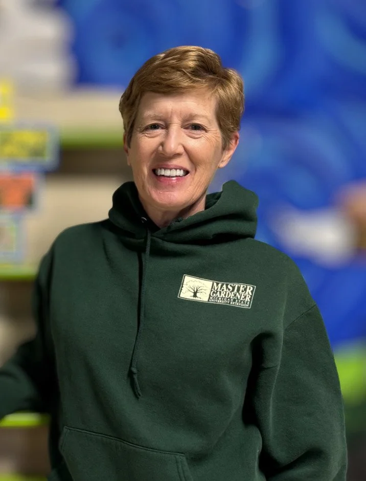 Smiling woman wearing a green hoodie with 'Master Gardener' logo inside a store with gardening supplies at Chelesea Senior Center.