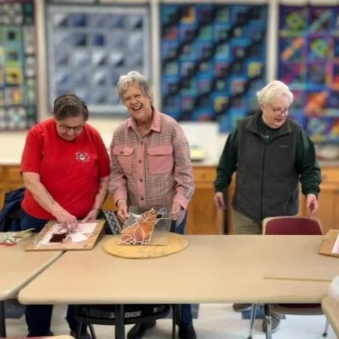 Three elderly women are standing around a table in a craft room, smiling and working on art projects with fabric and sewing supplies, with shelves of organized materials in the background.