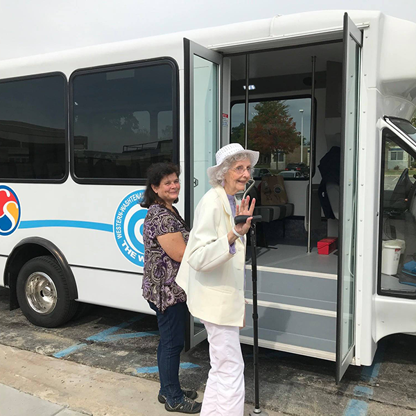 An elderly woman waving and smiling outside a white bus with a woman behind her, in a parking lot with handicapped parking space.