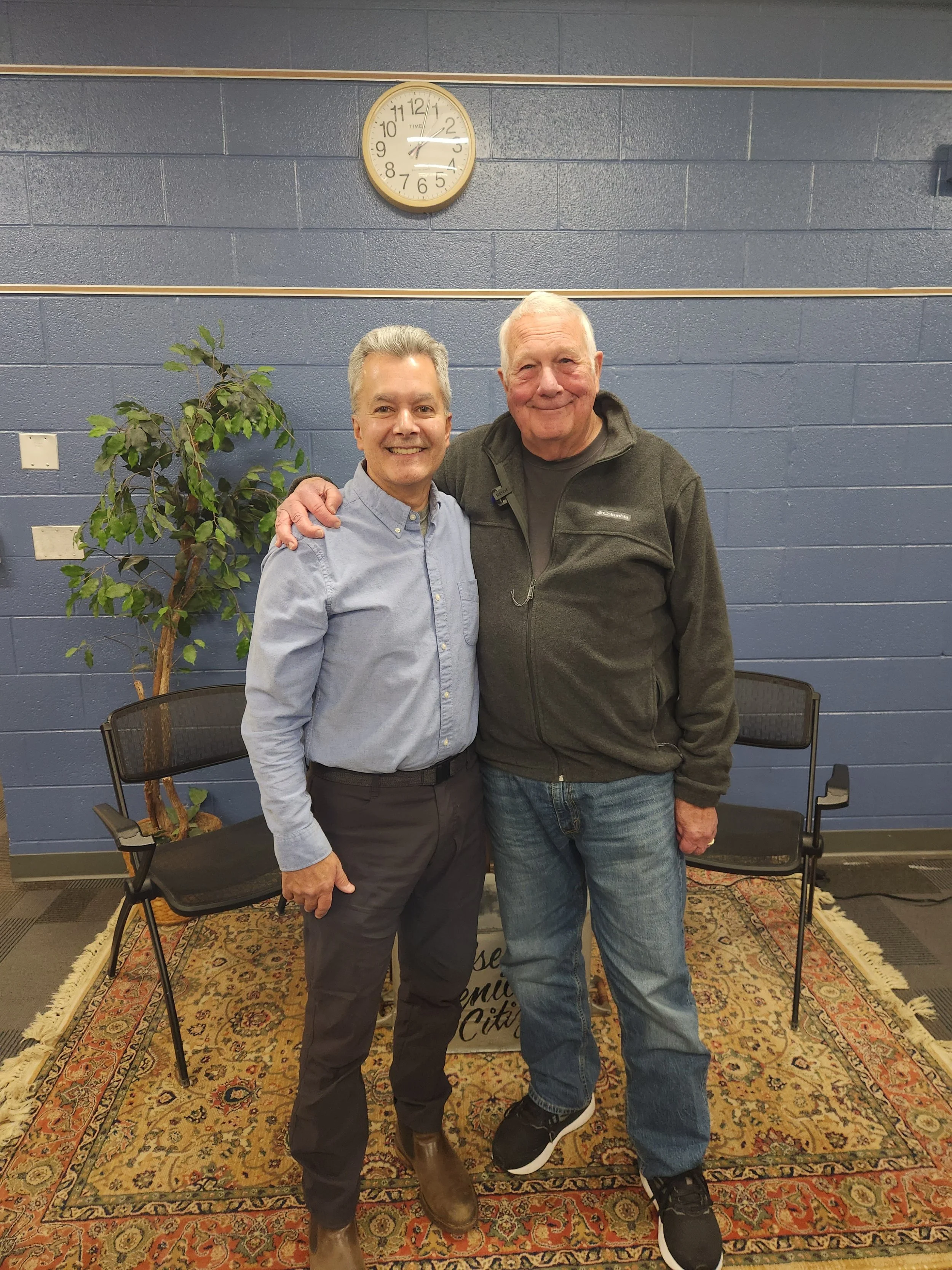 Two men standing together, smiling, with their arms around each other, in front of a blue wall with a clock. One man is wearing a light blue button-up shirt and dark pants, and the other is wearing a dark Columbia fleece jacket and jeans. There are black folding chairs and a patterned rug on the floor, with a plant to the left.