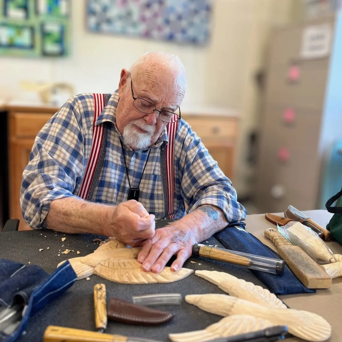 An elderly man with glasses and a white beard carving a wooden object at a worktable at Chelsea Senior Center, surrounded by carving tools and wood shavings in a workshop or craft room.