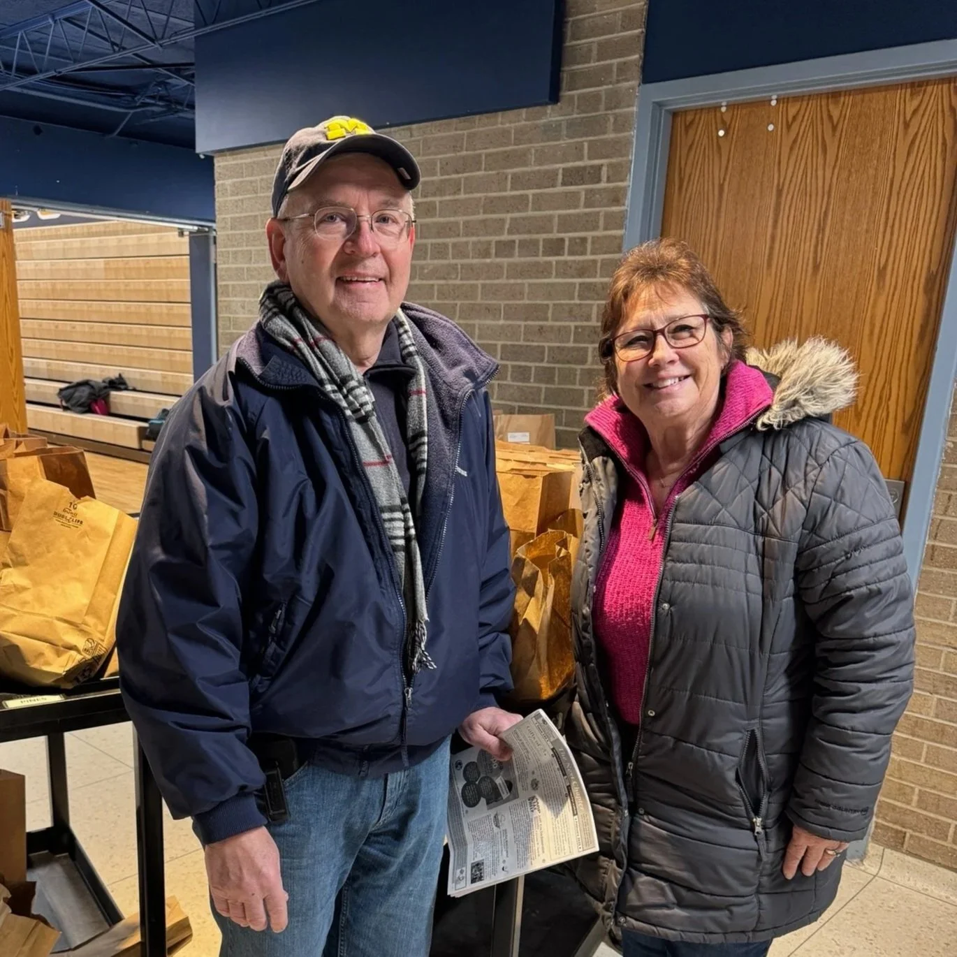 A man and a woman stand indoors, smiling at the camera. The man wears a baseball cap, glasses, a navy jacket, and a scarf. The woman wears glasses, a quilted gray jacket with a fur-lined hood, and a pink sweater underneath. Behind them are wooden and metal fixtures, and on the table in front of them are paper bags.