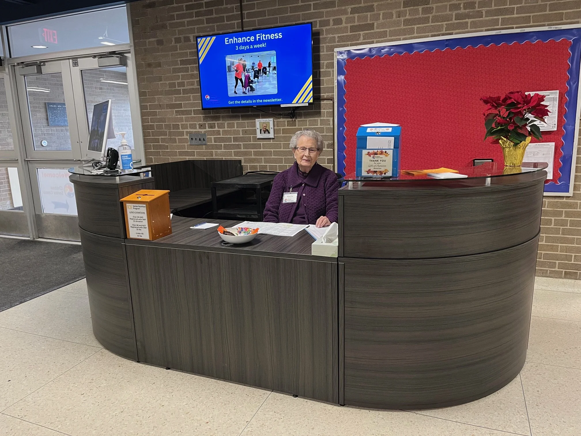 Hospital reception area with a curved dark wood desk, a senior woman seated behind it, a poinsettia plant in a yellow pot, a bowl of snacks, and a sign thanking donations. A bulletin board with red background and blue border, a screen advertising fitness classes, and a hand sanitizer pump near the glass door are visible.