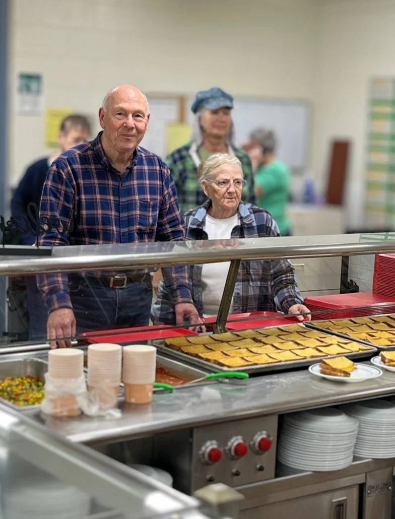 People serving food behind a cafeteria counter, with trays of food and disposable cups visible.
