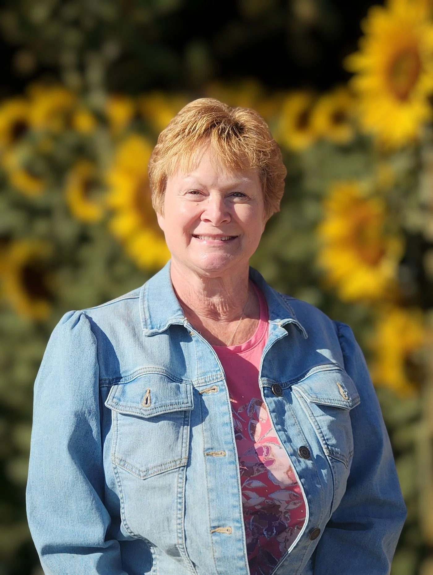 A smiling woman with short, reddish hair, wearing a denim jacket over a pink floral shirt, standing outdoors in front of a field of sunflowers at Chelesea Senior Center.
