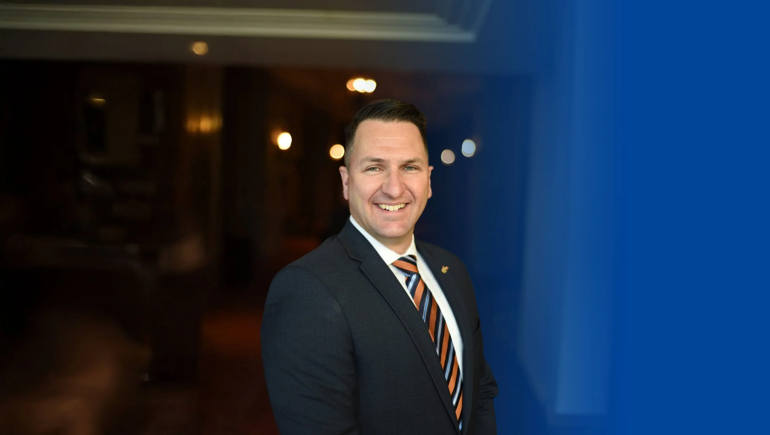 A man in a dark suit with a colorful striped tie smiling in an indoor setting with warm lighting.