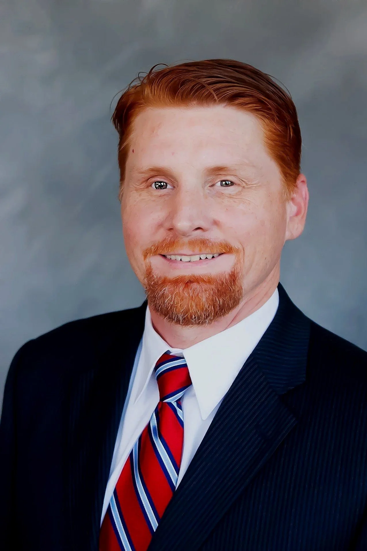 Headshot of a smiling man with red hair, a beard, wearing a dark suit, white shirt, and a red, white, and blue striped tie, against a gray background.