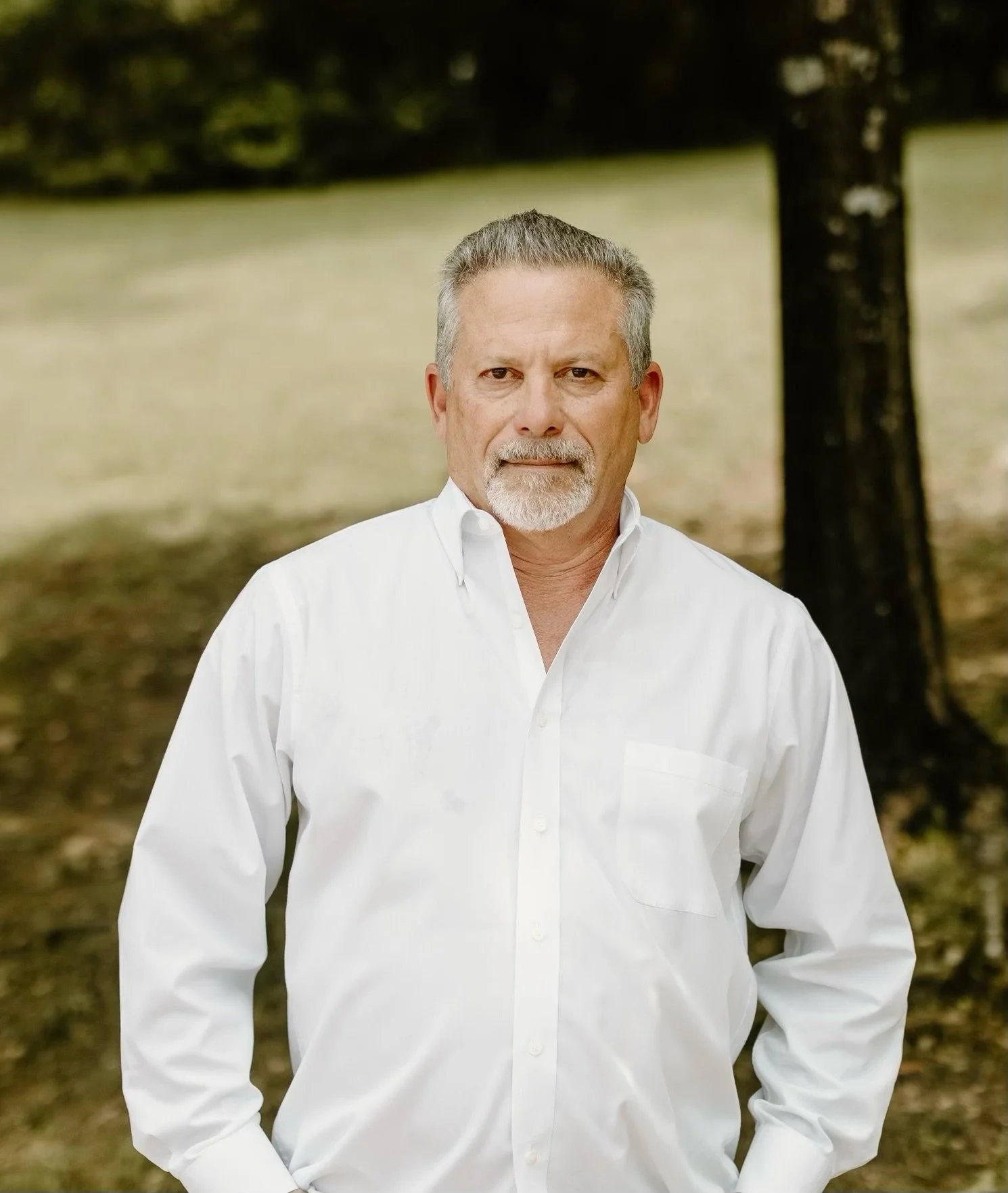 A middle-aged man with gray hair and a beard, wearing a white button-up shirt, stands outdoors in front of trees and a grassy area.