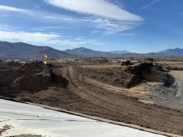 Construction site with excavator and dirt pile, mountains in the background under a blue sky.