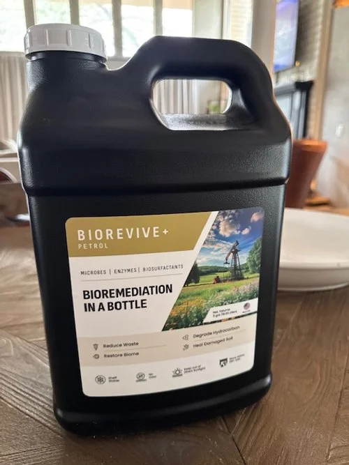 Black plastic jug with a white cap, labeled Biorevive+ Petrol, standing on a wooden table in a home setting.