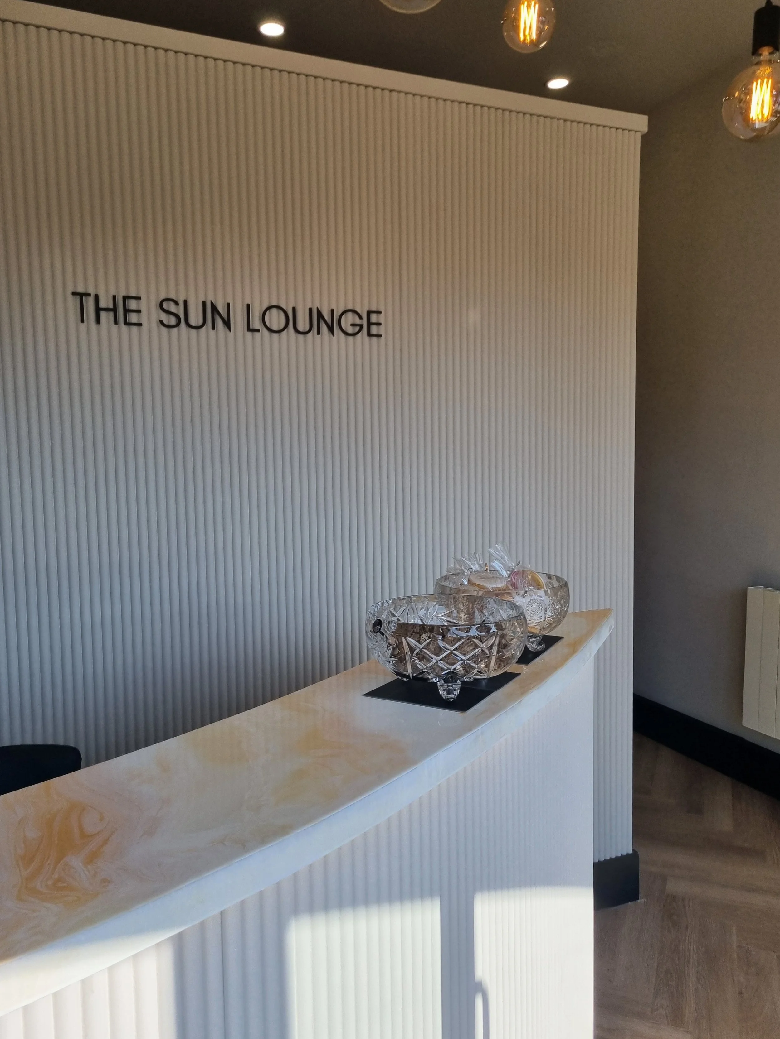 Decorative glass bowls on a white reception counter in the Sun Lounge, with a white textured wall and pendant lights in the background.