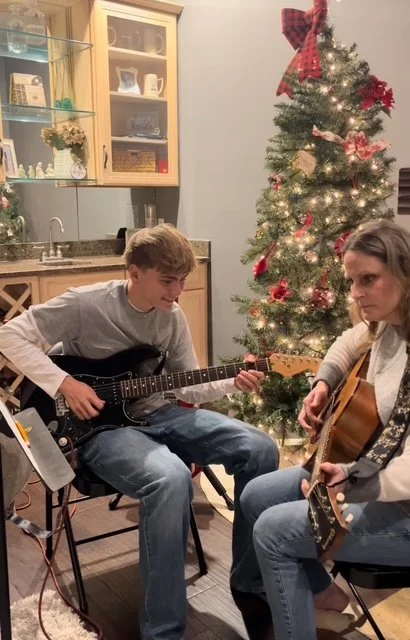 A young man and woman playing guitars together in a festive room with a decorated Christmas tree.