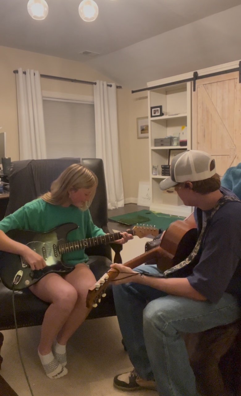 Two young people sitting in a living room playing electric and acoustic guitars.