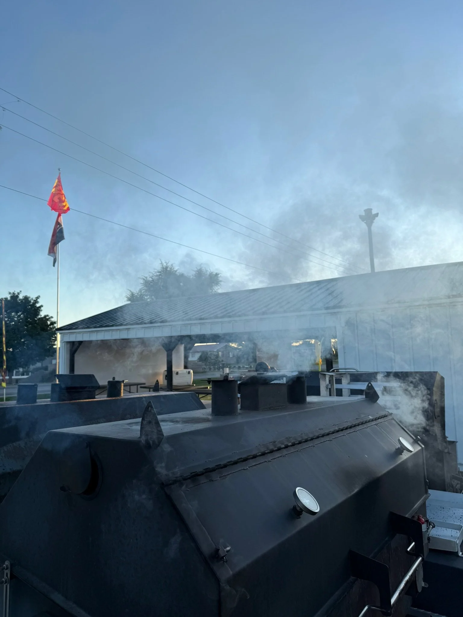 Two large black smoker grills with smoke rising from them are outdoors under a shelter with a gray metal roof. A flagpole with a red flag is visible to the left, and a cloudy blue sky is in the background.