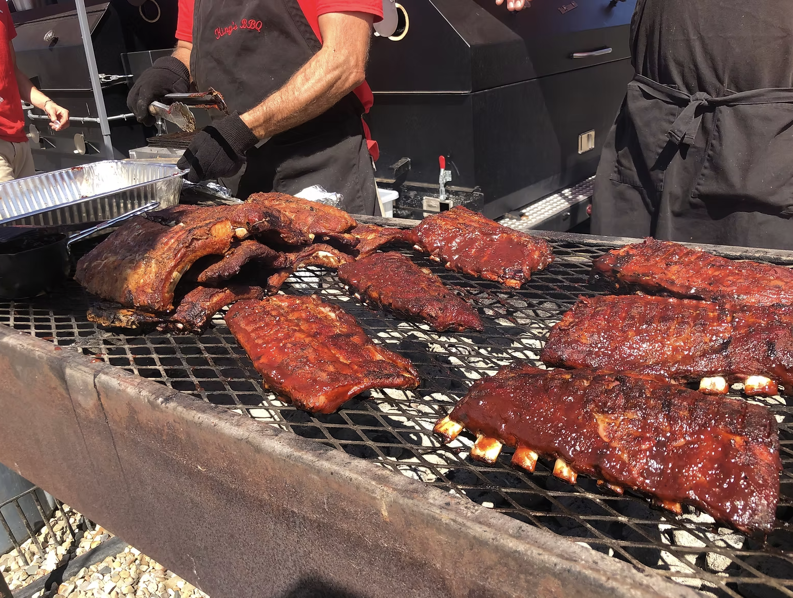 Barbecue grill with racks of ribs and pork on it, being cooked outdoors with people in black and red shirts in the background.