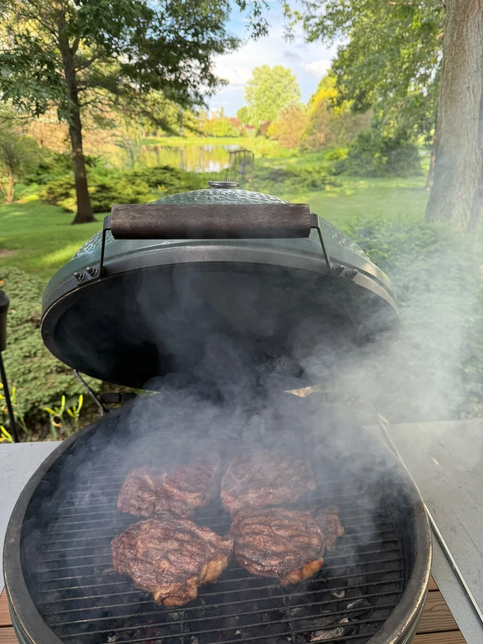 Close-up of a round black outdoor grill with a lid open, cooking three steaks with smoke rising, set on a wooden table outdoors in a lush green park with trees, grass, and a pond in the background.