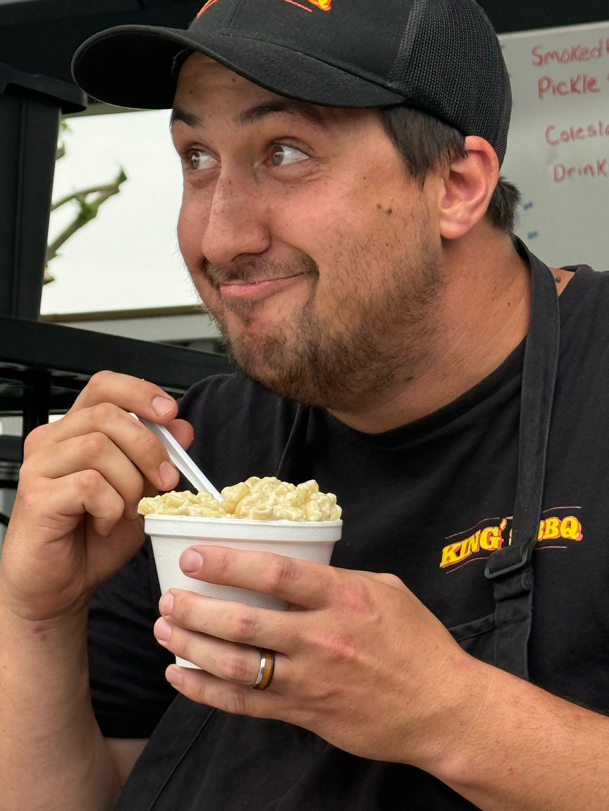 A man with a beard and a black baseball cap smiling while holding a cup of macaroni and cheese, with a whiteboard in the background that has written menu items.