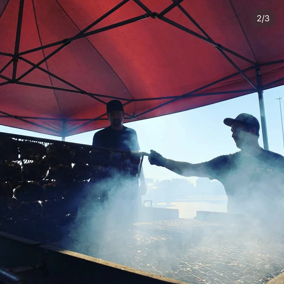 Two men grilling food under a red canopy with smoke rising from the grill.