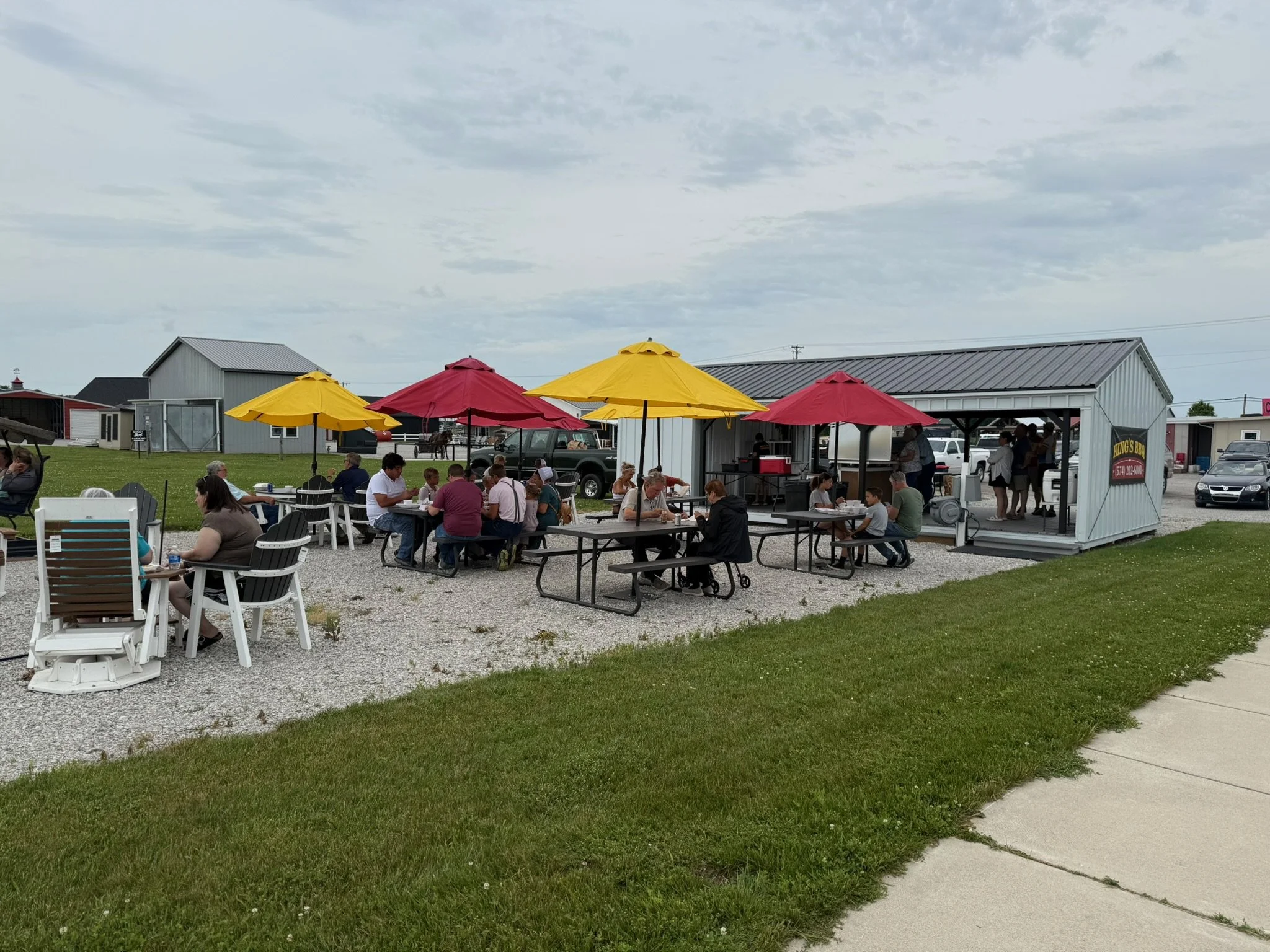 Outdoor dining area with people sitting at tables under yellow and red umbrellas at a casual eatery, metal building with a sign, parked cars, cloudy sky, green grass, and a concrete sidewalk.