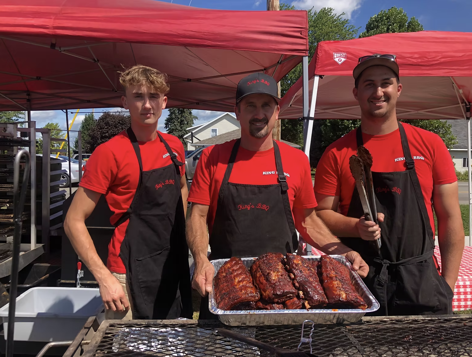 Three men at a barbecue stand, wearing red shirts and black aprons, with smoked ribs on a tray in front of them. They are outdoors under red tents, with trees and houses in the background.