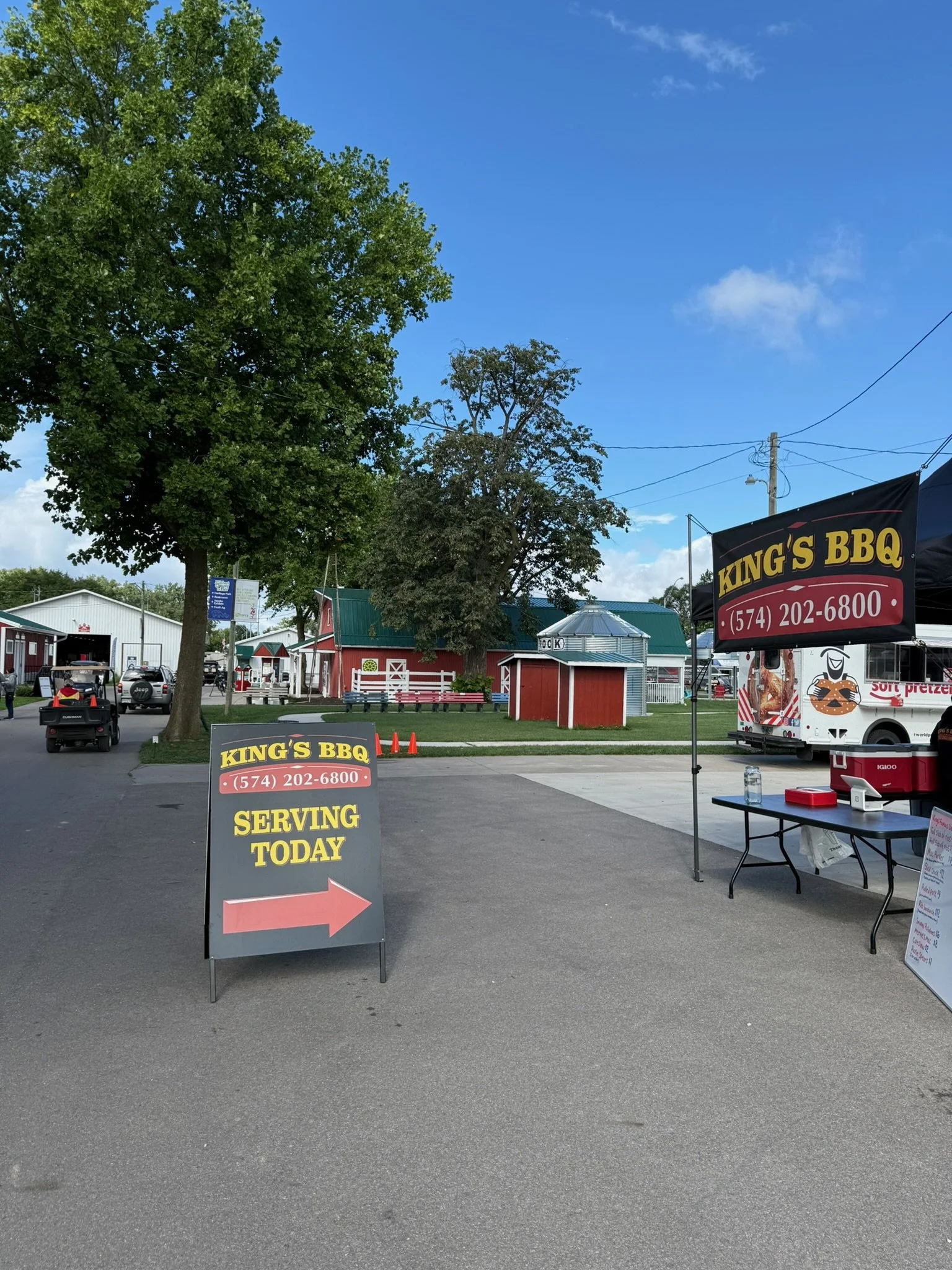 A street scene with a large tree, a red barn, and a food stand with signs for King's BBQ. The signs indicate the food stand is serving today and provide a phone number.