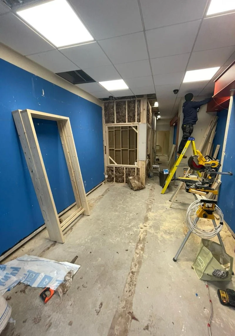 Construction site inside a room with partially installed walls, a worker on a ladder working on the ceiling or wall, construction tools and materials scattered around, blue wall on the left side, and exposed framing at the back.