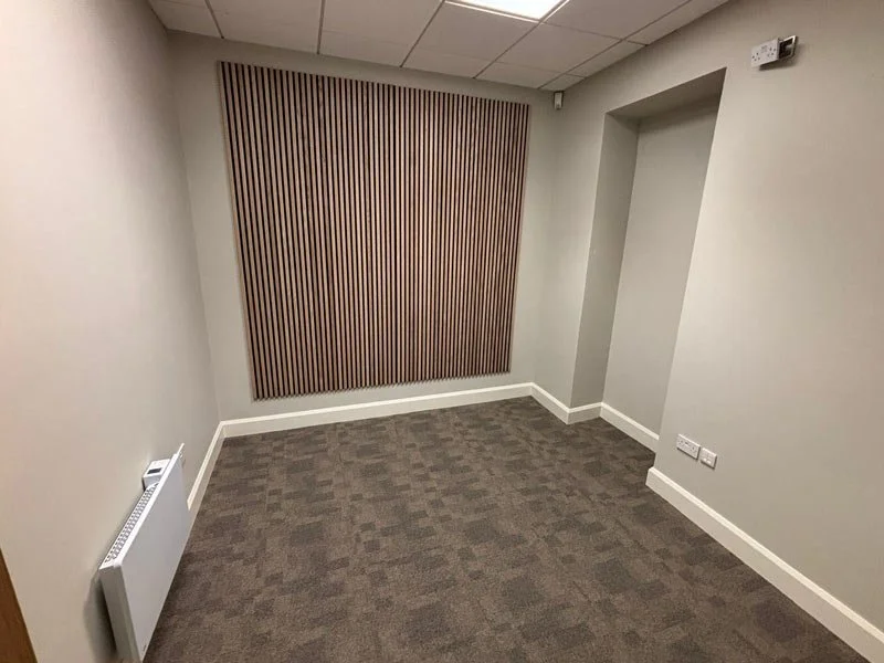 Empty room with beige carpet, light grey walls, and wooden paneling on the back wall. There is a white radiator on the left wall and electrical outlets on the right wall near the bottom. The ceiling has a suspended tile ceiling with a visible vent.