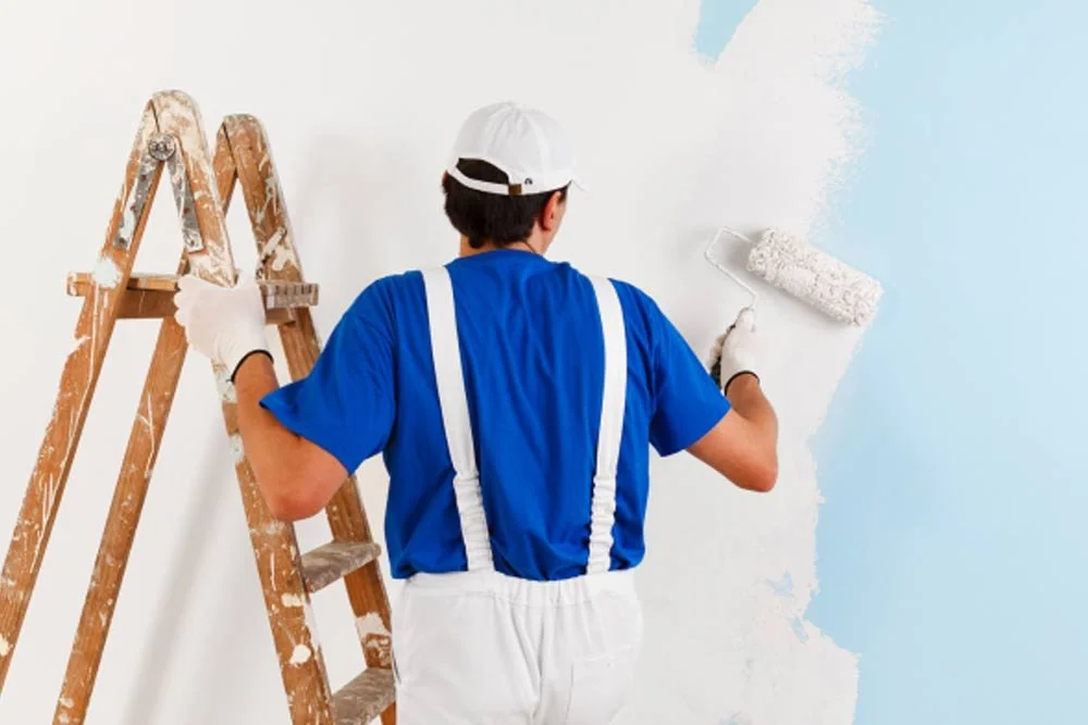 Man painting a wall white using a paint roller, standing next to a wooden ladder.