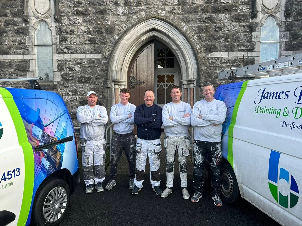 Five painters in work clothes posing with their arms crossed, standing in front of a stone church building with two painting vans on either side.
