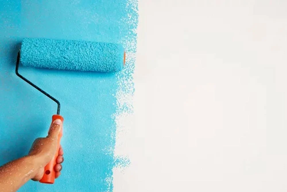 Close-up of a hand using a paint roller to paint a wall light blue.