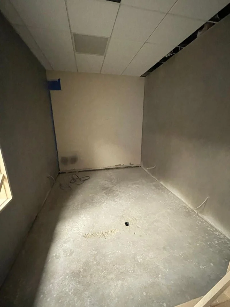 An unfinished room with drywall walls, a concrete floor, and a ceiling with some ceiling tiles and open space for additional tiles. There is a small window on the left wall, and some wires are visible on the floor.