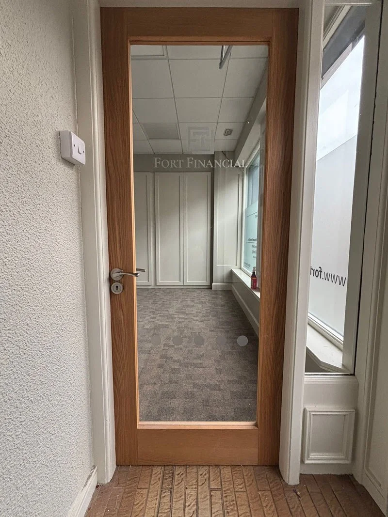 An interior view through a wooden-framed glass door into an empty room with white walls, a window, and a gray carpeted floor.