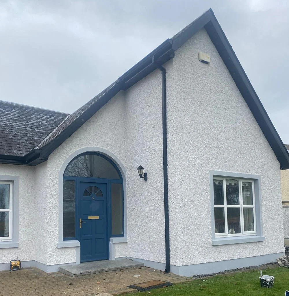White house with blue door and window frames, black rain gutter, and a lamp on exterior wall, with a small front yard and cloudy sky.
