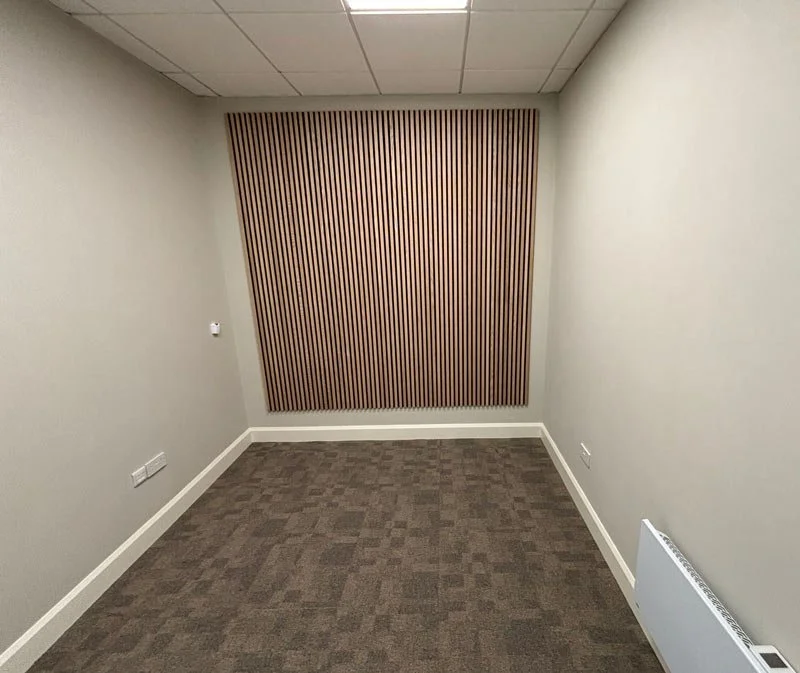 Empty small office room with gray walls, a textured wood accent wall, gray carpet, and white baseboards.