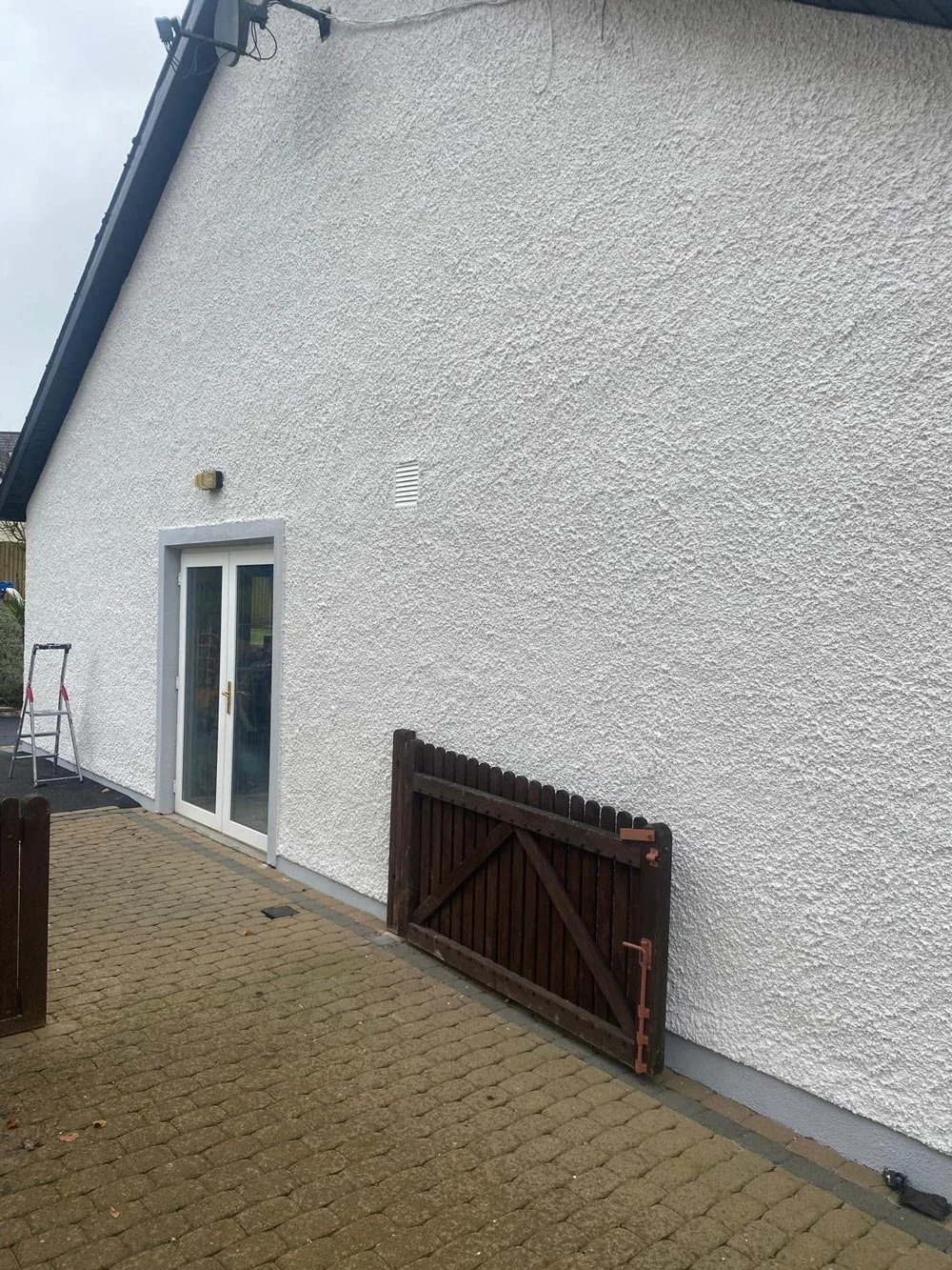A white residential house wall with a sliding glass door, a small outdoor step ladder, a wooden fence gate, and a paved patio area.
