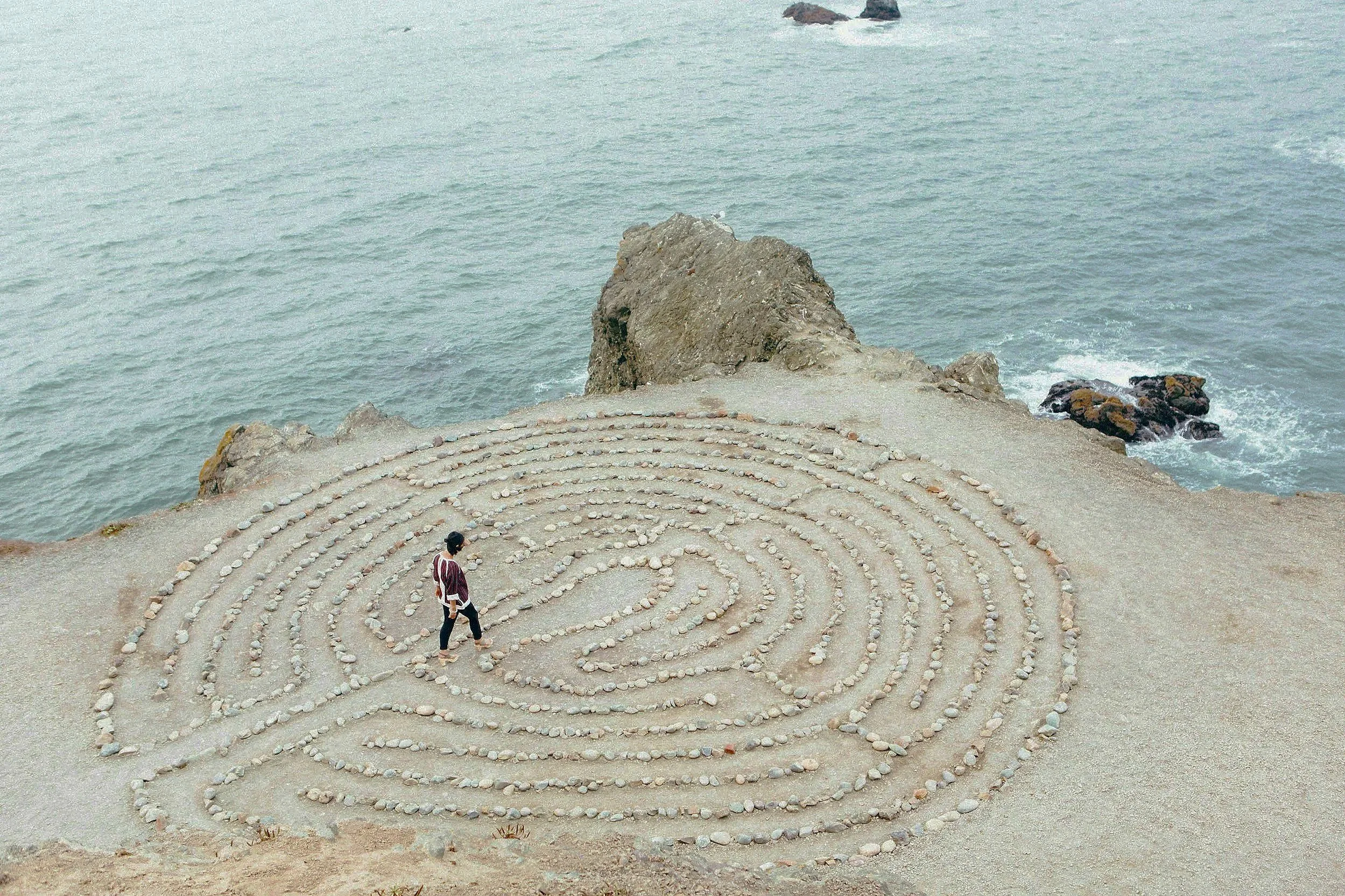 A person walking along a gravel pattern on a beach, with concentric circles made of small stones, near a rocky shoreline with ocean waves.