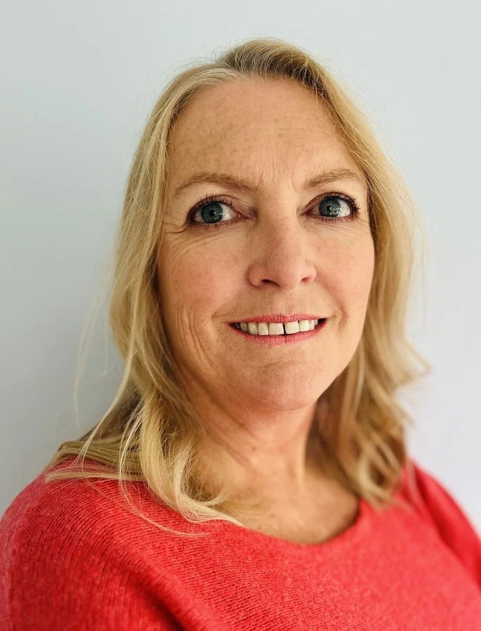 A woman with blonde hair, blue eyes, and fair skin smiling at the camera, wearing a red top, against a plain light-colored background.