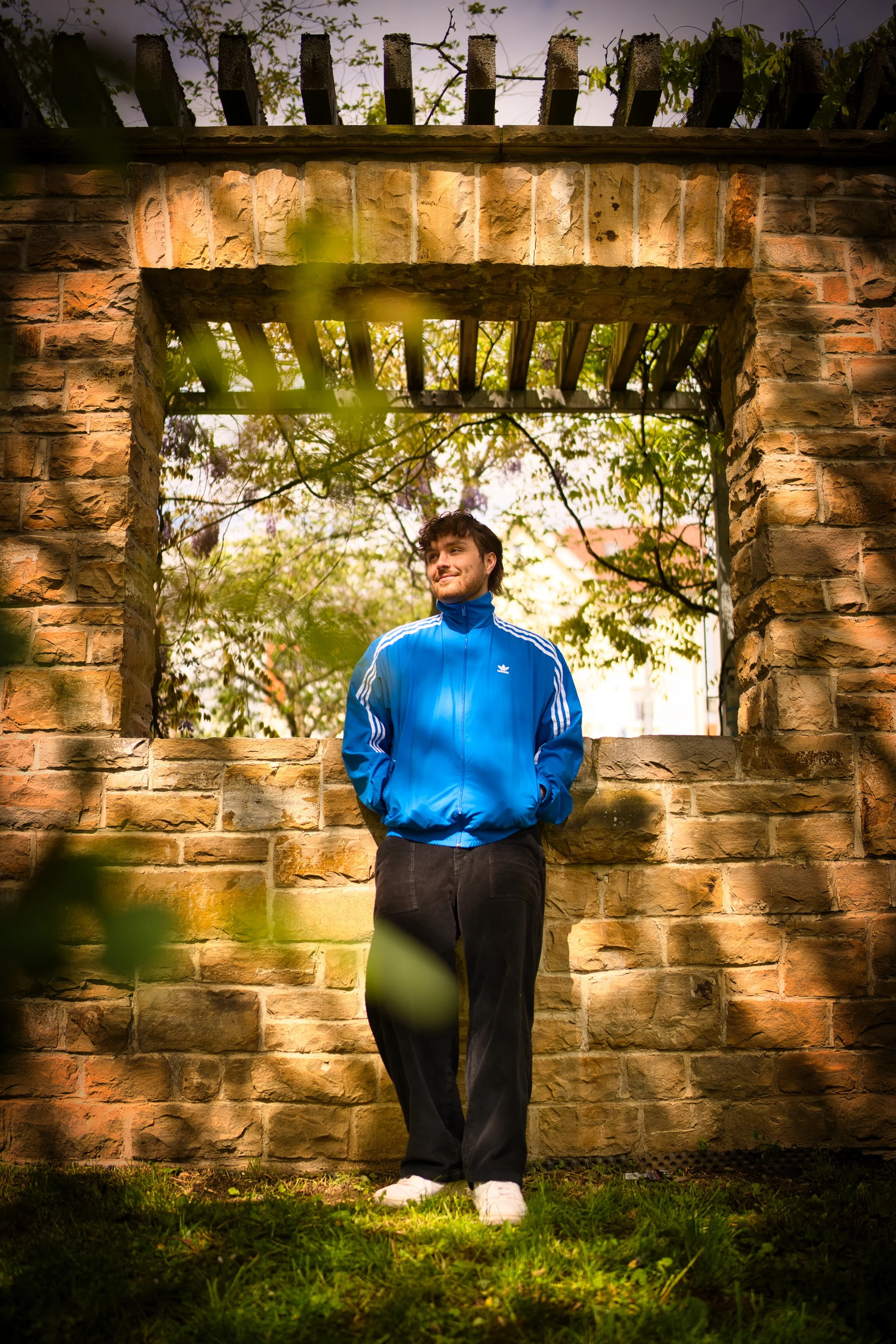 A young man in a bright blue Adidas jacket and black pants stands with hands in pockets in front of a brick wall and window, with trees and sky behind him.