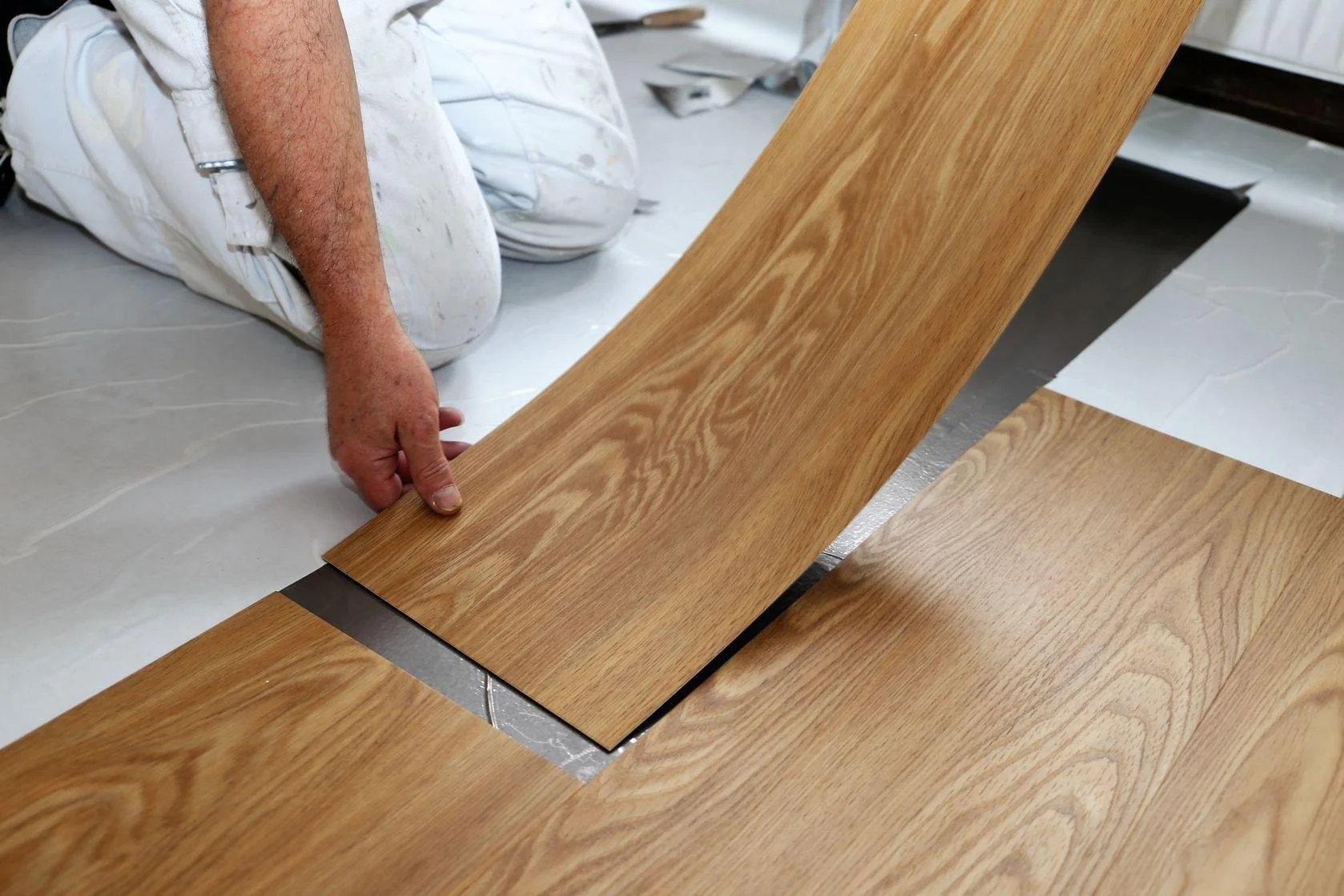 A person installing or laying wooden flooring on a surface, kneeling down and holding a piece of wood.