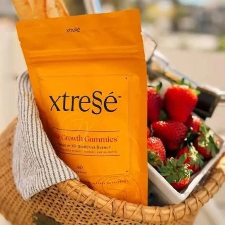 A person holding a yellow pack of Xtresé Growth Gummies in a hand wearing a gray glove, with a basket of strawberries in the background.