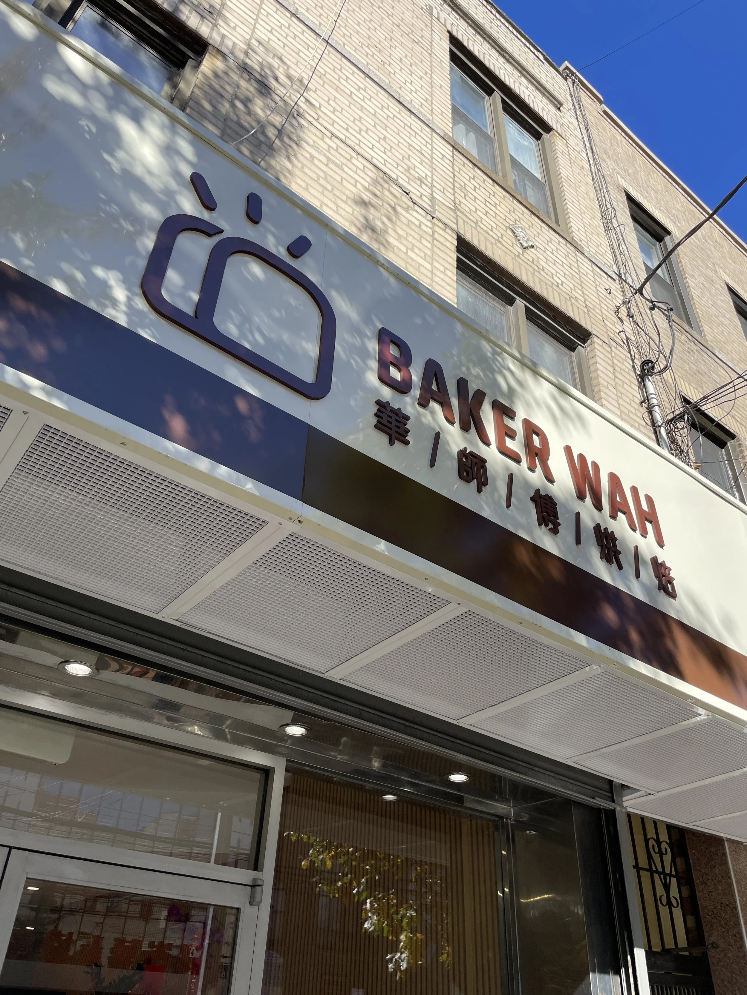 Storefront sign for Baker Wah with English and Chinese text, part of a building with a brick exterior and windows, under a blue sky.