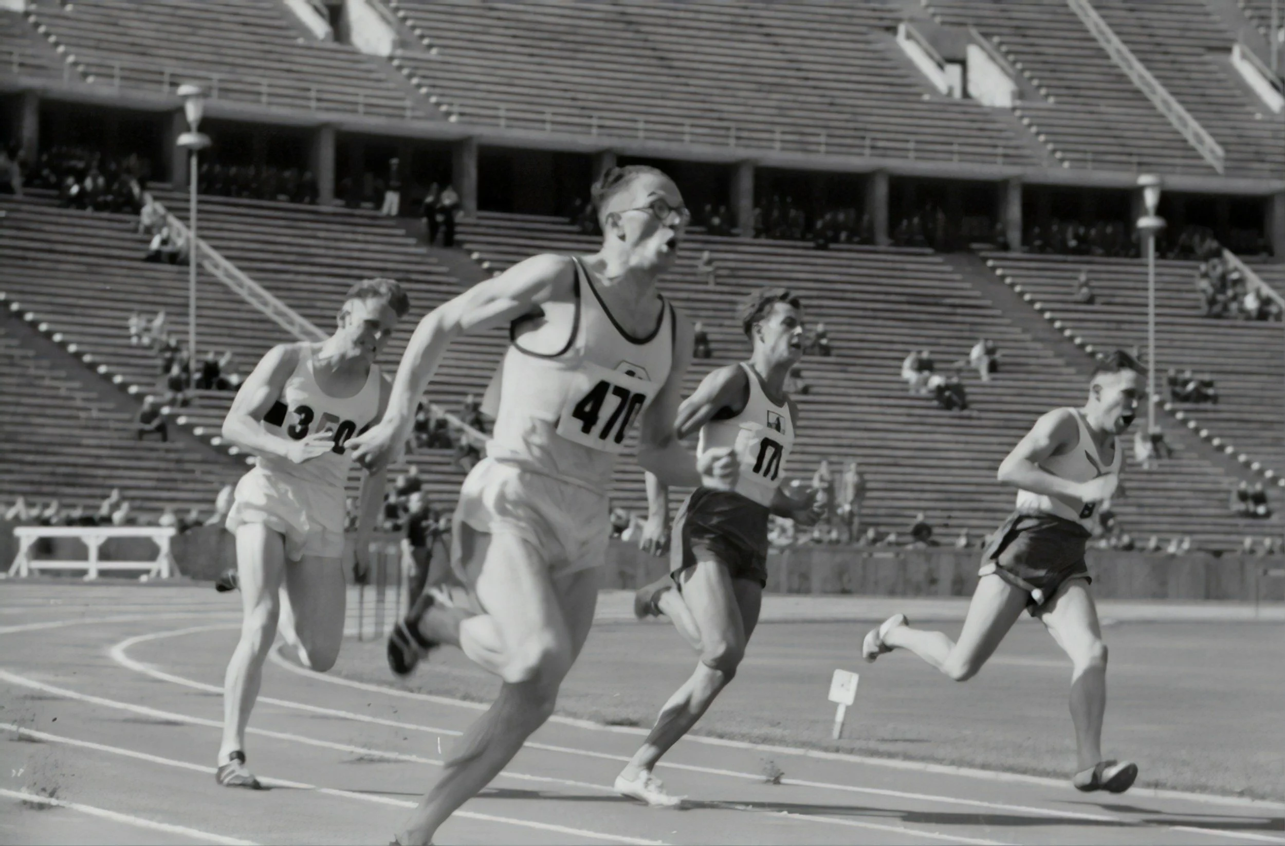 Four male athletes running on a track during a race at a stadium with mostly empty seats.