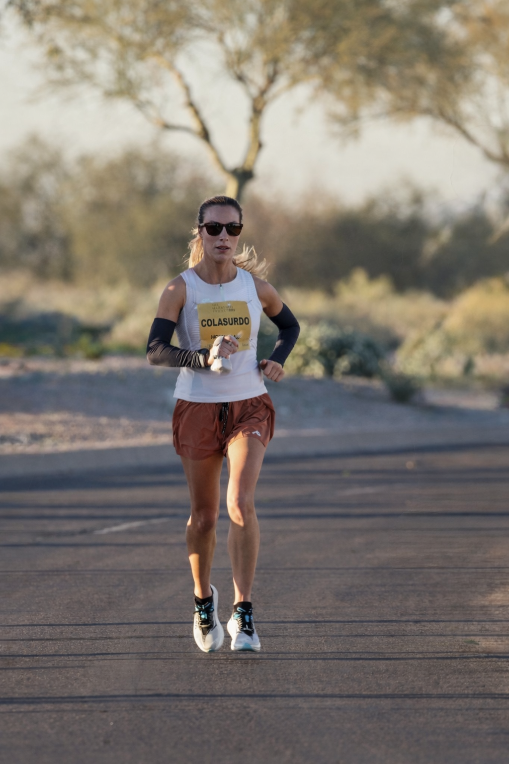 A woman running outdoors on a paved path during daytime, wearing sunglasses, a white sleeveless shirt, copper-colored shorts, and black arm sleeves, with a race bib labeled 'COLASURDO' pinned to her shirt.