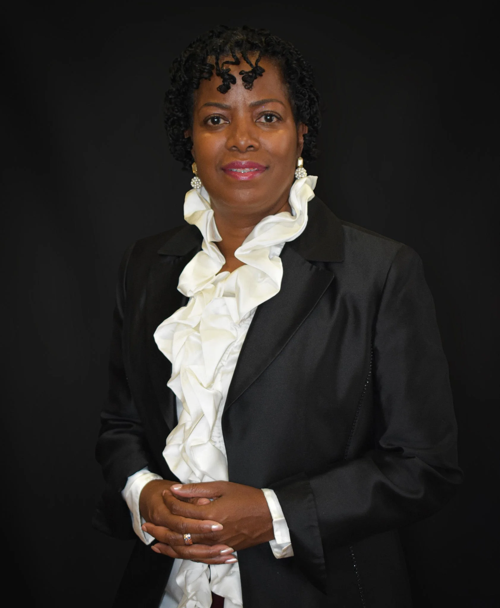 A woman with short, curly black hair, wearing a black blazer over a white ruffled blouse, standing against a plain black background. She has earrings and a ring on her left hand.
